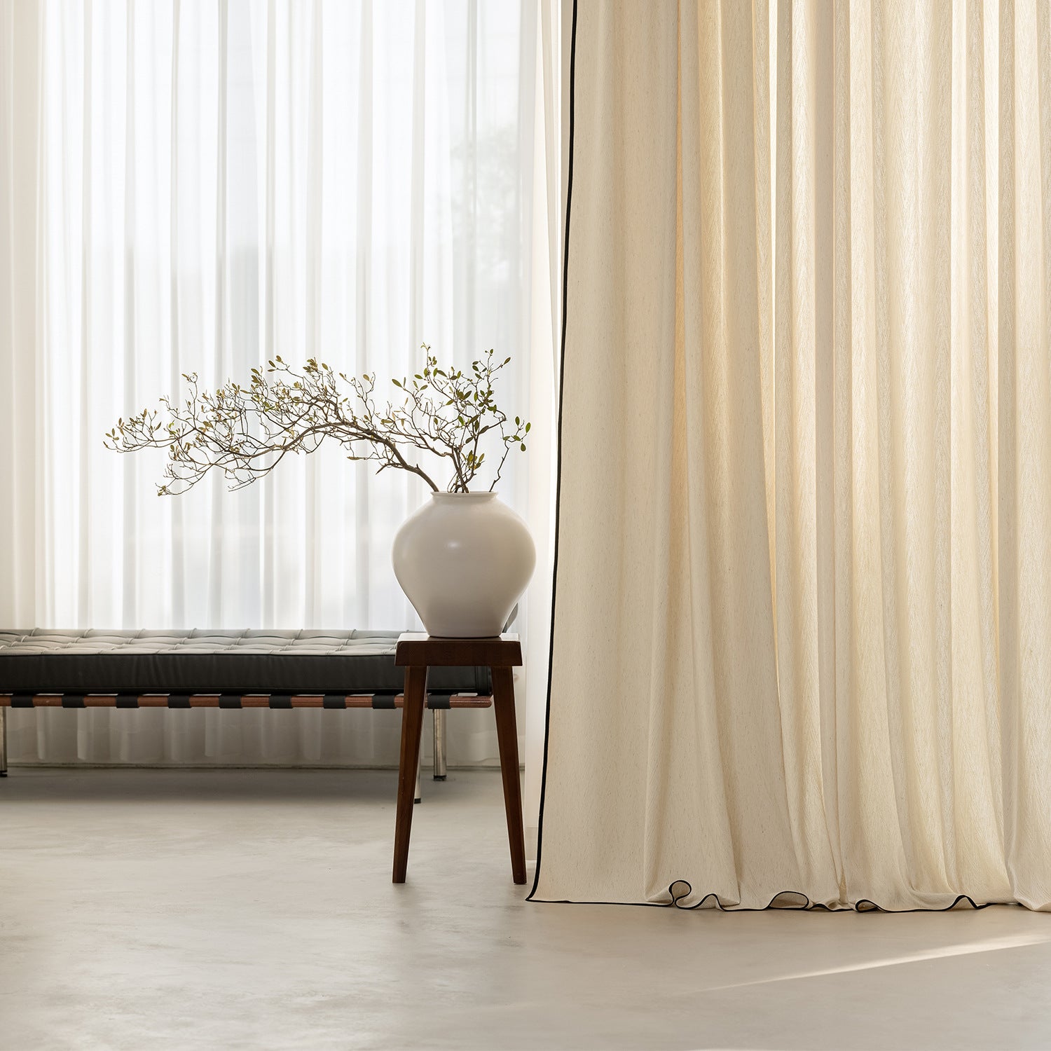 White curtain with a vase and branches on a wooden stool in a room.